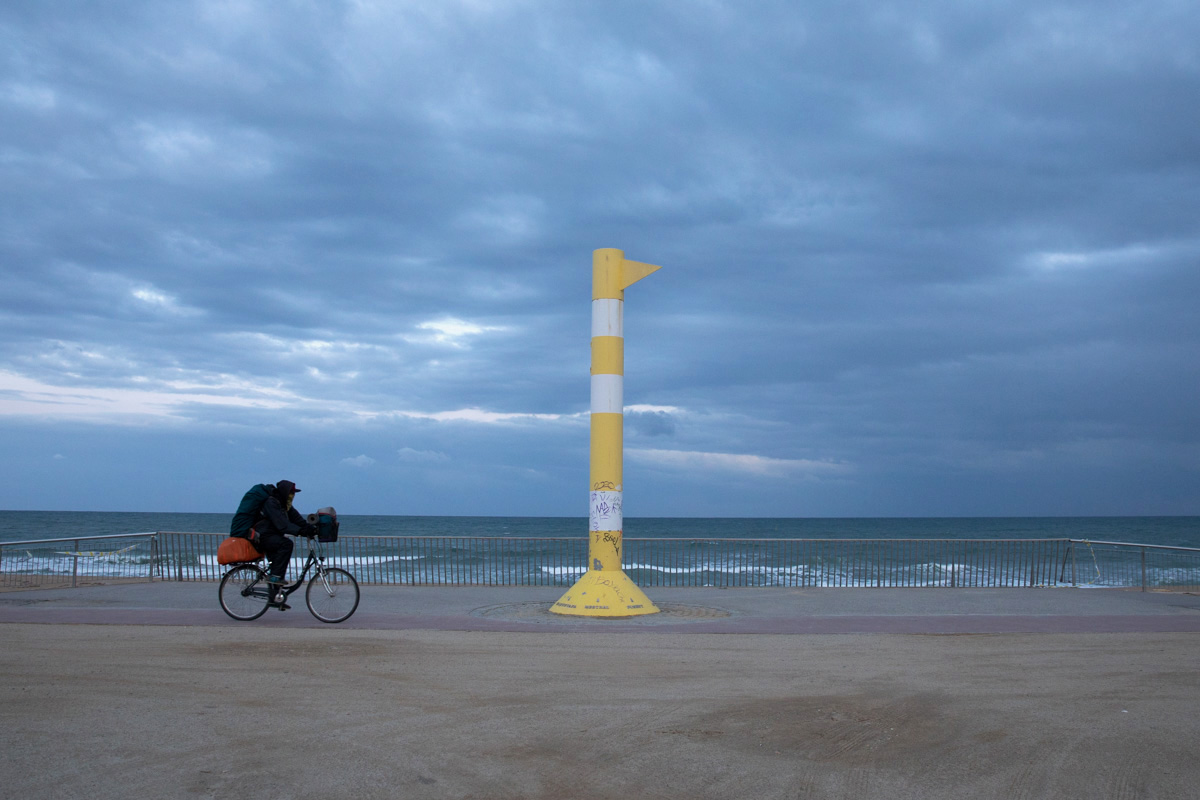 A young man from Venezuela is driving along Bogatell’s beach promenade at 6:00 in the afternoon through a deserted Barcelona, due to the confinement decreed by the State of Alarm caused by the Coronavirus crisis. In his backpack, he carries a sleeping bag, a tent and a cooking stove. Before confinement he used to work as a circus artist. He does not want to go into one of the shelters settled up by the City Council for confinement because he fears social panic. Barcelona, March 26. // Un joven de Venezuela está circulando en bicicleta por el paseo marítimo de la playa del Bogatell a las 6 de la tarde, por una Barcelona desértica debido al confinamiento decretado por la crisis del coronavirus. En su mochila lleva un saco de dormir, una tienda de campaña y un hornillo. Antes de la cuarentena trabajaba como artista de circo. No quiere ir a al albergue establecido por el Ayuntamiento de Barcelona porque teme el pánico social y las multitudes. Barcelona, 26 de marzo. Foto: Eva Parey.